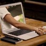 Laptop montrant un bras, main tendue, table en bois, smartphone, stylo, tasse de café