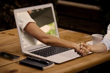 Laptop montrant un bras, main tendue, table en bois, smartphone, stylo, tasse de café