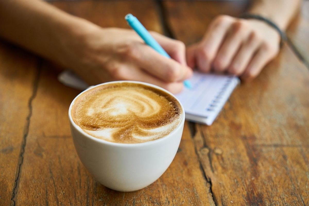 Une tasse de café au lait avec mousse une main écrivant dans un carnet Une tasse de café au lait avec mousse une main écrivant dans un carnet